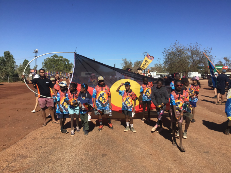 Luke with a group of young children smiling while holding the Aboriginal flag 