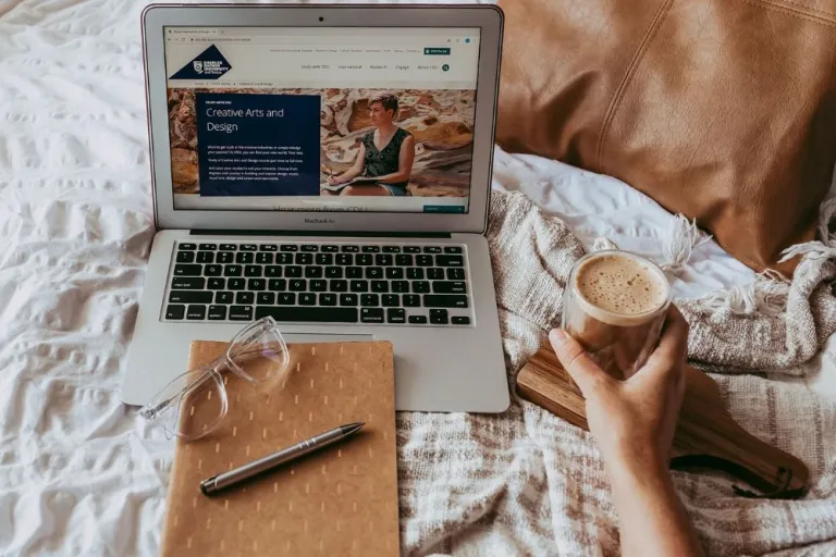 Study space on a bed featuring a laptop, notepad, glasses, pen and a coffee in hand