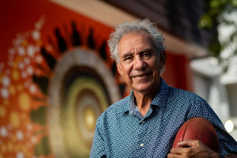 An Aboriginal man with greying hair and a warm smile holds a football while looking at the camera. He is standing in front of a First Nations artwork.