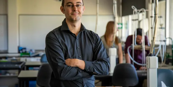 Dr Dylan Irvine standing in front of a bench in a laboratory facing the camera, with electrical cables, and people out of focus in background