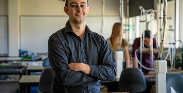 Dr Dylan Irvine standing in front of a bench in a laboratory facing the camera, with electrical cables, and people out of focus in background