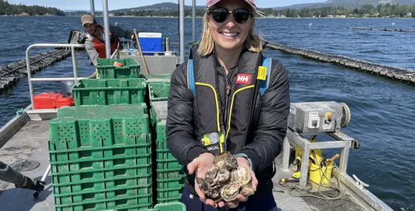 Samantha Nowland, wearing sunglasses and a cap, standing on a boat on water, holding a quantity of oysters in both hands. A person is in the background operating the boat.