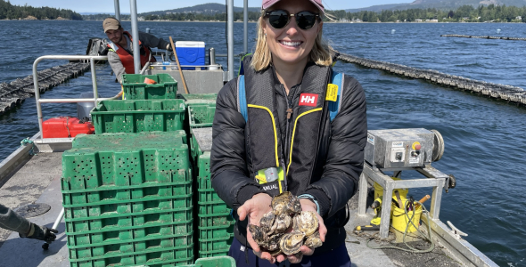 Samantha Nowland, wearing sunglasses and a cap, standing on a boat on water, holding a quantity of oysters in both hands. A person is in the background operating the boat.