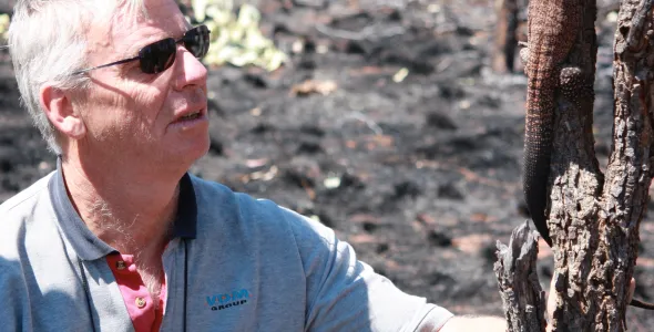 Dr Noel Preece, wearing sunglasses, with binoculars hanging round his neck, looking at a lizard on a burnt tree trunk, surrounded by burnt ground