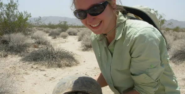 Dr Chava Weitzman wearing sunglasses holding a tortoise, in arid-looking country with small shrubs in the background