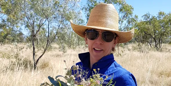 Dr Donna Lewis head and shoulders, wearing hat and sunglasses and holding a bunch of native flowers and leaves, with grass and trees in background
