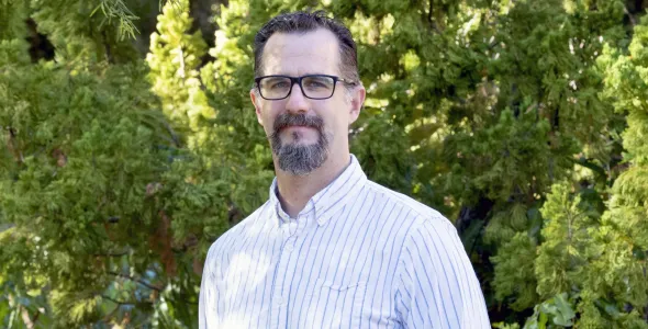 Dr Keller Kopf with a beard and glasses, wearing a shirt with vertical light blue stripes, with dense green foliage behind