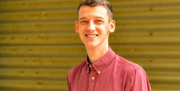 Sam Williams - young man smiling and wearing a light red button up shirt smiling in front of corrugated iron wall