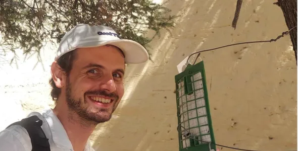 Andrew Coates, head and chest, on the left of the photo, in a white shirt and a white cap, smiling at the camera, in front of a white wall, with tree branches above his head. A bug trap attached by wires to a tree trunk which is just visible at the right of the image