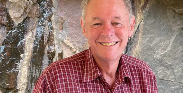 Steven Tickell, head and chest, wearing a red shirt with white cross hatching, with a rock face behind him