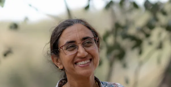 Kamaljit Sangha, head and shoulders, wearing a patterned top and glasses, with a blurry green and brown background