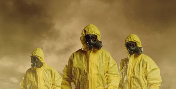 three people wearing full-body yellow hazmat suits and black gas masks. They are standing in a smoky, dusty, or foggy environment with a dark brownish sky, giving the scene an intense, possibly hazardous or post-apocalyptic feel. Yellow caution tape runs across the lower part of the image, suggesting a restricted or contaminated area.