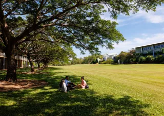 large expanse of grass lawn with buildings on far side, three people sitting on grass in shade of large trees; more buildings visible behind trees