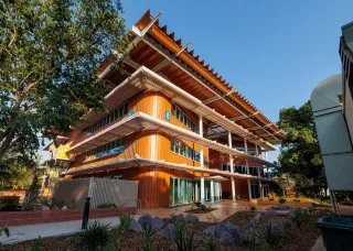 A contemporary three-storey university building with bright orange cladding, wide glass windows, and layered white verandas. The structure features striking angled roof panels for shade, with a blue number 8 sign marking the building. The foreground shows landscaped garden beds with rocks and native plants, and a clear blue sky forms the backdrop.