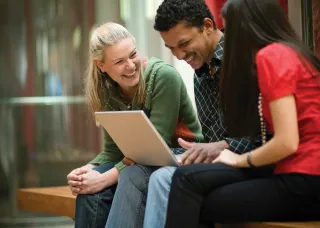 Three young adults sit on a bench indoors, smiling and laughing while looking at a laptop together.