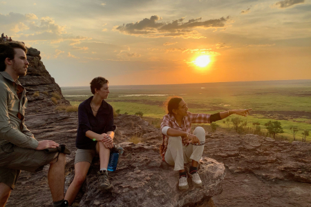 three people on a rock high above a background plain with sun low in sky
