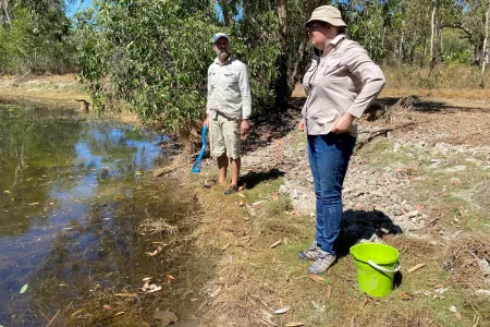 Two people standing on the bank of a water body with a green bucket in the foreground and trees in the background