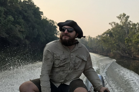 Cameron Baker wearing sunglasses and hat, steering a boat on a river lined with forest, with wake behind