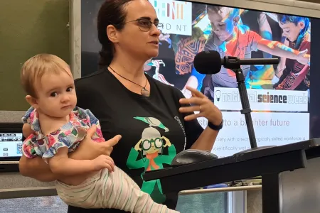 Dr Carla Eisemberg speaking into a microphone at a lectern, holding a baby on one arm