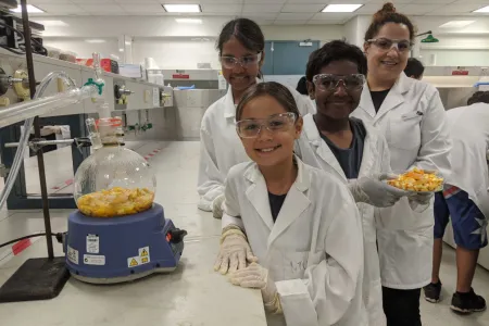 three children and an adult wearing white coats and protective glasses, in a laboratory