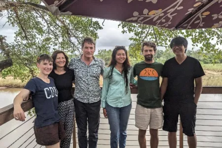 six people standing in a row, posing for photo, with vegetation in background