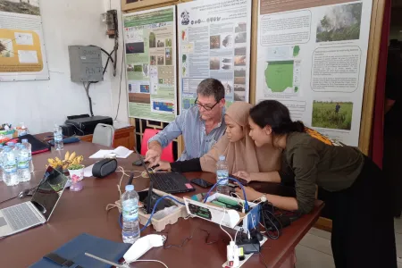Three people looking at a laptop on a table with electronic equipment. Academic posters on wall in background