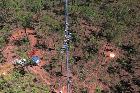 Aerial view of a tall tower with two people working on it. Trees, vehicles, and solar panels far below