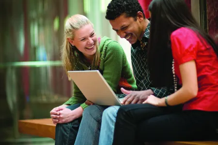 Three people sitting on a bench. Person in middle looking at laptop screen.