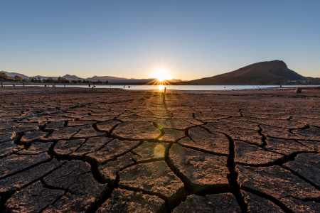 Cracking soil of a dry lake bed, with water in the distance and hills behind. Sun low in the sky behind hills