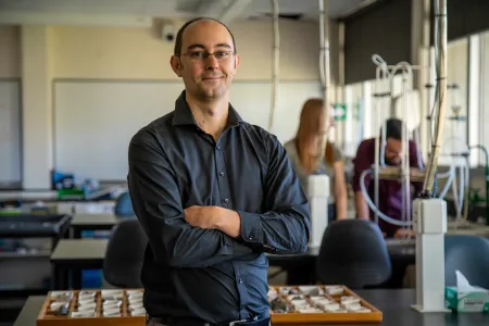Dr Dylan Irvine standing in front of a bench in a laboratory facing the camera, with electrical cables, and people out of focus in background