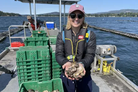 Samantha Nowland, wearing sunglasses and a cap, standing on a boat on water, holding a quantity of oysters in both hands. A person is in the background operating the boat.