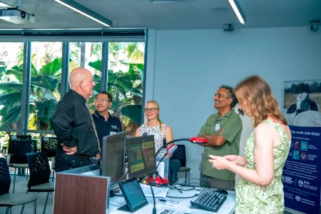 Five people stand chatting near lecture room lectern, with large palm fronds visible through windows in the background