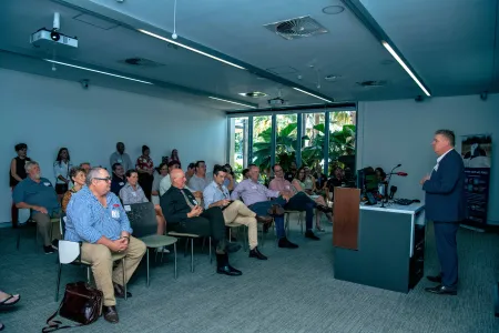 Man wearing suit standing at lectern in front of room full of people sitting on chairs. Through windows in background are seen several palms with large fronds, close to the windows.