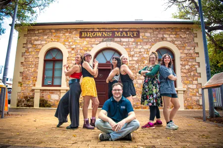 Six women stand behind one man who is seated on the floor while the women look strong and proud standing in poses akin to Charles Angel's.