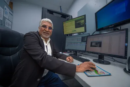 Man sitting at desk with computer screens in front of him. He is holding the mouse but looking directly at the camera and smiling. 