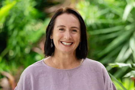 Caucasian women with brown hair smiles widely at the camera. She is outside in front of some green foliage and is wearing a lilac coloured top. 