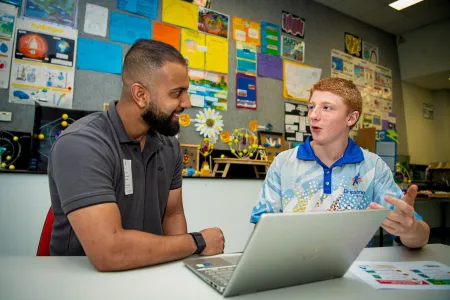 Two people sitting at a desk. A laptop is open in the middle. The University student on the left smiles at the Year 9 student on the right. They're in an animated discussion. 