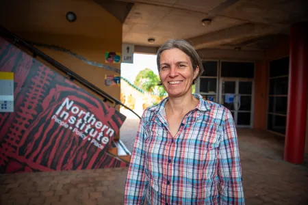 Caucasian woman with grey hair and a plaid blouse standing in front of a stairway that says Northern Institute. 