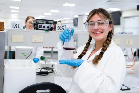 A young woman wears a white lab coat and is holding a glass beaker inside a lab. She has a wide smile. 
