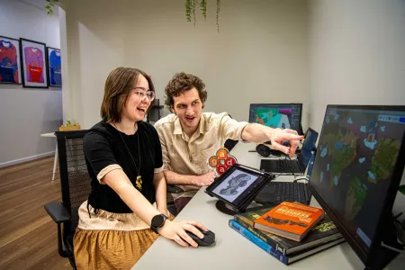 Young woman and man sit at a desk looking animatedly at a computer screen. The young man is pointing. The young woman has a bright smile on her face. 