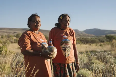 Two First Nations women stand outside surrounded by grass and mountains. One is holding a clay pot with drawings on it. They both look out into the distance.