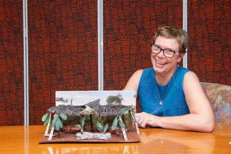A Caucasian woman sits smiling at a table. She is wearing a blue sleeveless dress. She has one of her architectural design models in front of her.