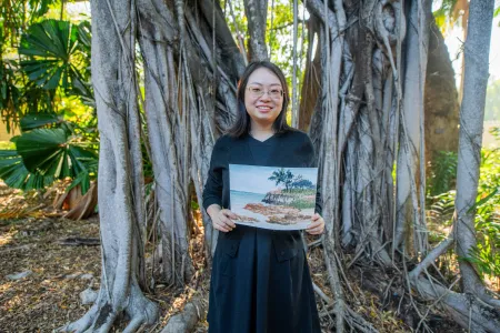 Young woman stands in front of native Australian trees on the Casuarina Campus in Darwin. She wears a black dress and cardigan and holds one of her watercolour pieces.