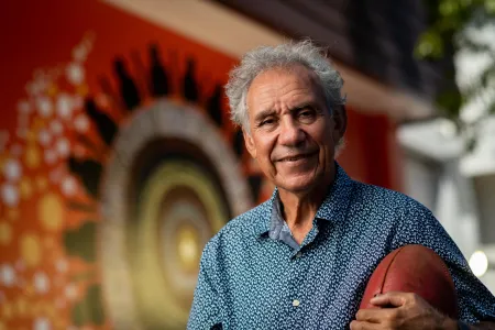 An Aboriginal man with greying hair and a warm smile holds a football while looking at the camera. He is standing in front of a First Nations artwork.