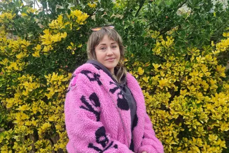 Young woman sits in front of wattle tree with yellow flowering. She wears a pink fluffy coat. 