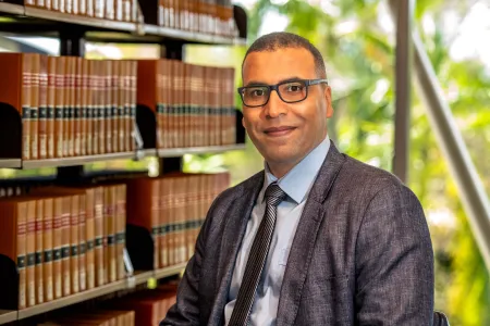 A man wearing glasses, a grey suit jacket, light blue shirt, and striped tie sits in a library with shelves of books behind him. He is smiling slightly and looking at the camera, with greenery visible through the window in the background.