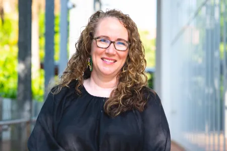 A woman with long curly brown hair and glasses is smiling outdoors. She is wearing a loose black blouse and colorful earrings, standing in a bright walkway with greenery and tall pillars in the background.