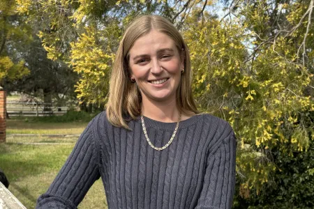 Young Caucasian woman standing outdoors looking at camera with a smile. She is wearing a grey jumper. 