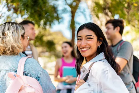 A young woman with brown hair smiles at the camera. She is wearing a white collared shirt. Next to her is a group of people engaged in discussion. They are standing outside. 
