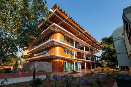 A contemporary three-storey university building with bright orange cladding, wide glass windows, and layered white verandas. The structure features striking angled roof panels for shade, with a blue number 8 sign marking the building. The foreground shows landscaped garden beds with rocks and native plants, and a clear blue sky forms the backdrop.